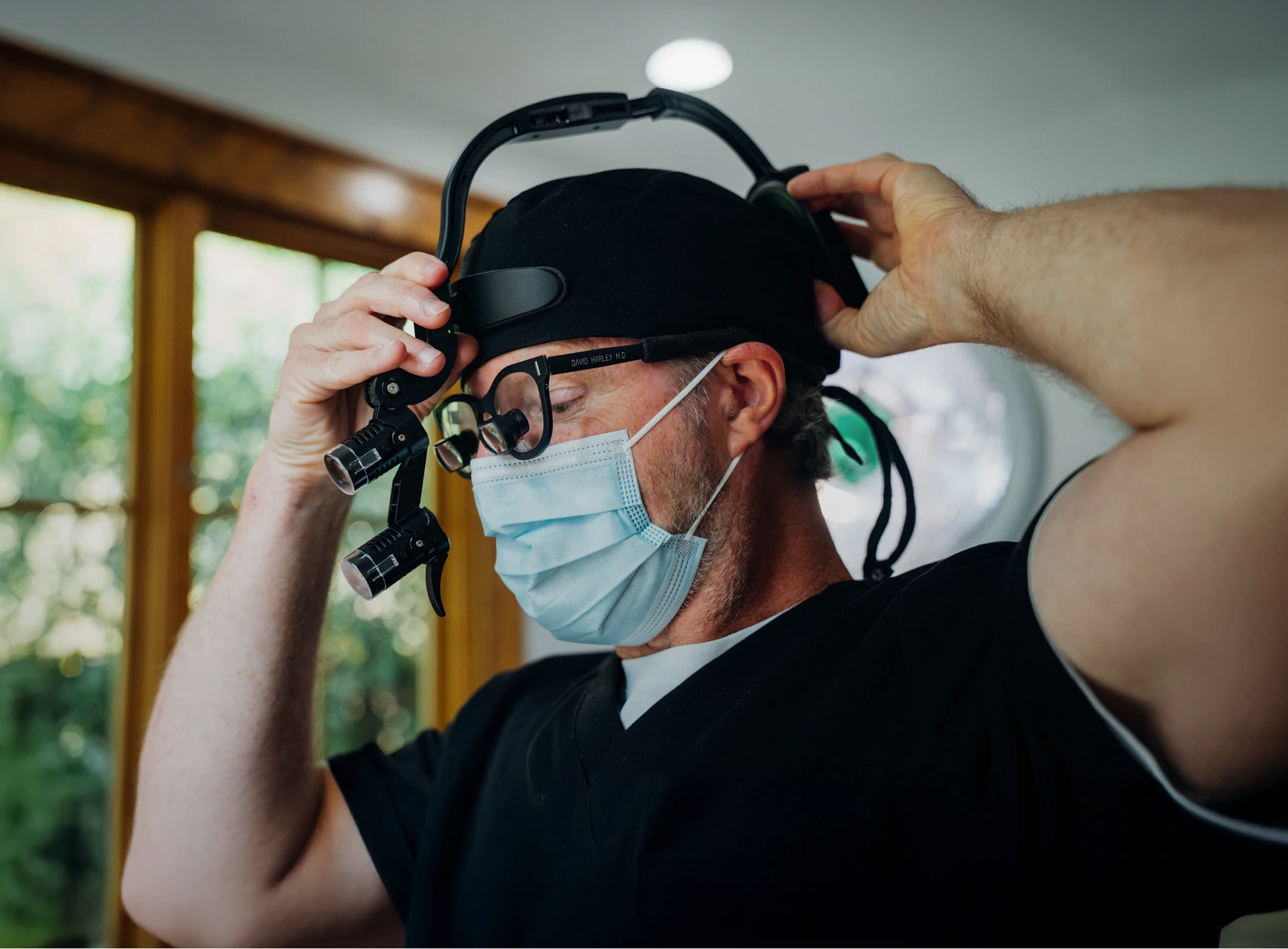 Dr. Harley, wearing a black surgical cap and face mask, adjusts a pair of specialized surgical loupes with integrated lighting on his head.