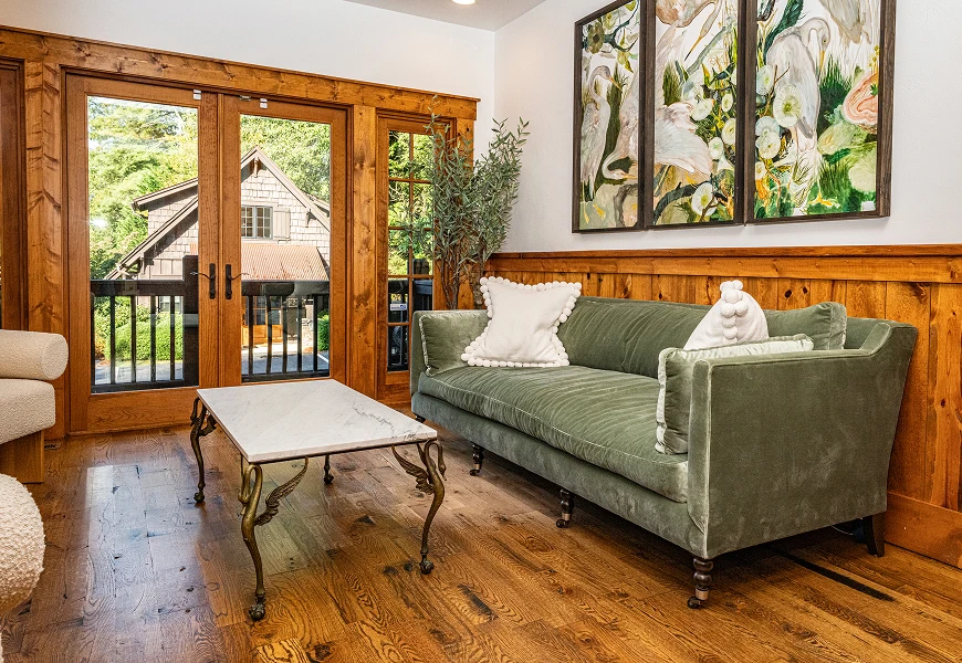 The interior of a waiting area featuring a sage green velvet sofa with white pom-pom pillows, a marble-topped coffee table with ornate legs, and glass French doors leading to an outdoor deck.