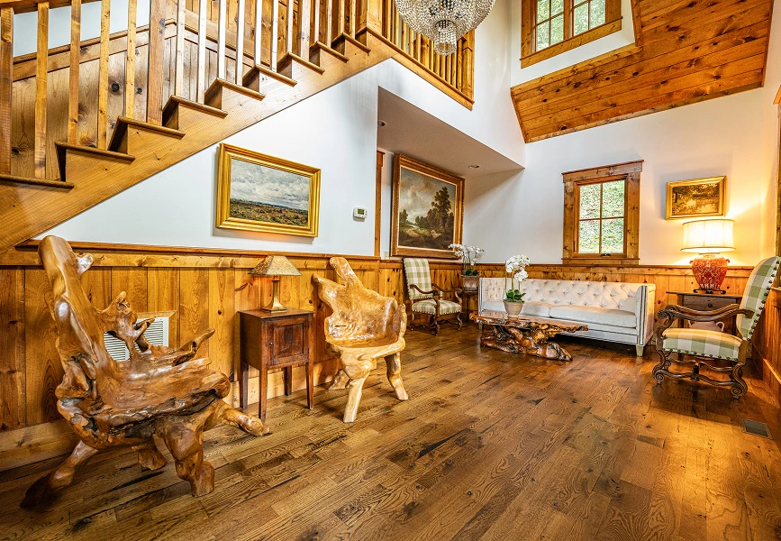 A spacious room with vaulted ceilings and wood paneling, featuring unique hand-carved wooden chairs, a white tufted sofa, and a staircase with a wooden banister.