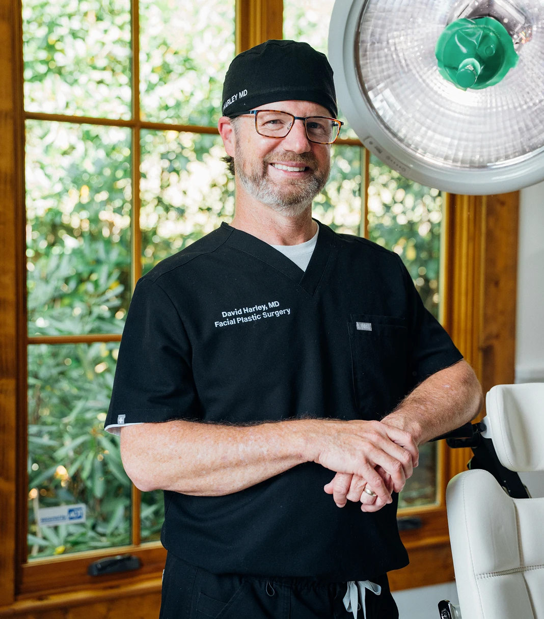 Dr. Harley stands in a clinical room wearing black scrubs embroidered with "David Harley, MD Facial Plastic Surgery," smiling with his hands clasped in front of a large window and a surgical light.