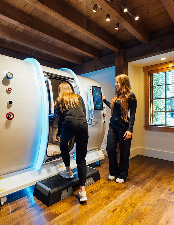 Two women in black attire stand near a large, futuristic white hyperbaric oxygen chamber with glowing blue LED trim inside a rustic room with exposed wood ceiling beams.
