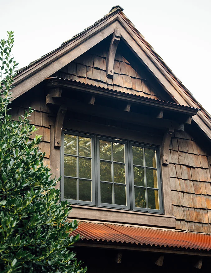 A vertical architectural shot of a rustic, dark wood-shingled building featuring a large multi-pane window under a gabled roof and a corrugated metal awning.