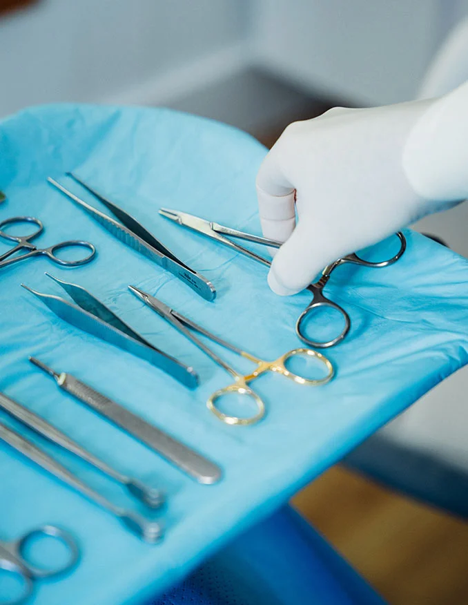 A close-up of a hand in a white sterile glove selecting a surgical instrument from a tray of metal tools laid out on a blue sterile drape.